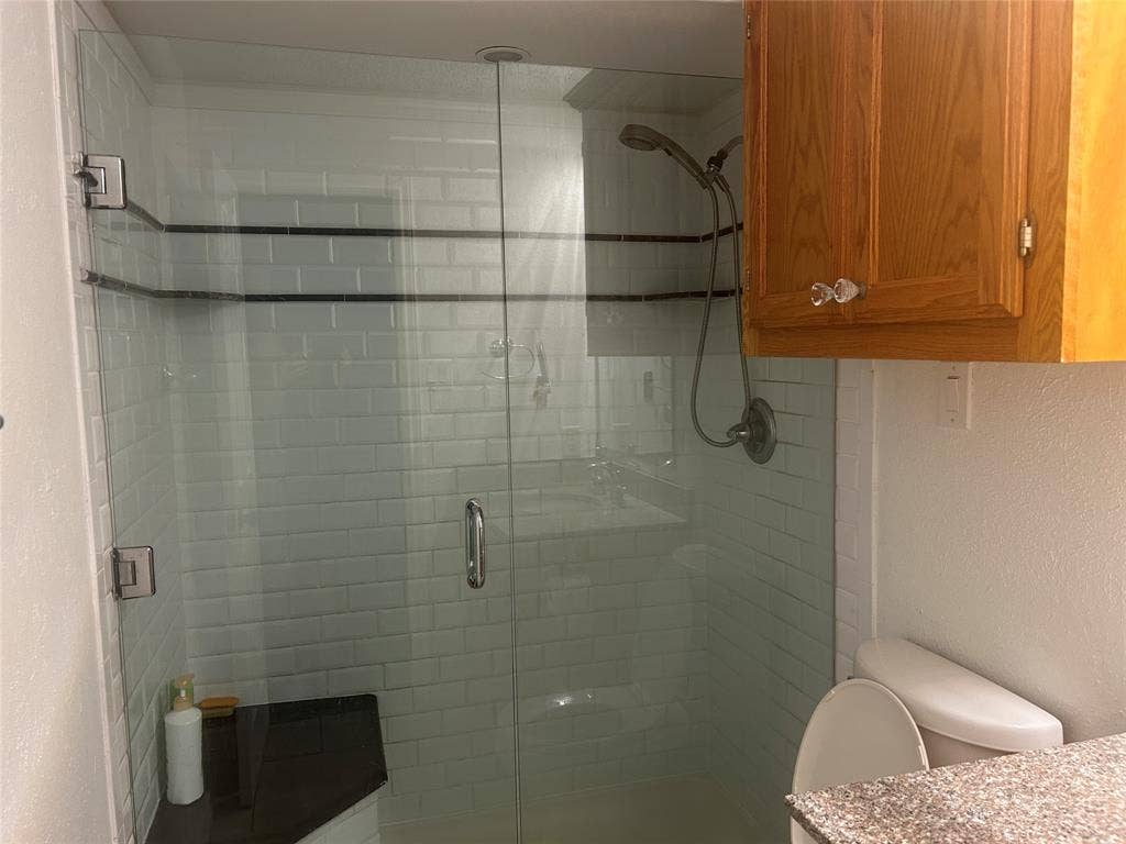 1314 Frio Lane, Garland, TX 75040 Walk-in shower featuring a frameless glass door, white subway tile surround with dark accent banding, built-in corner bench, handheld shower head, and wood-finish cabinetry