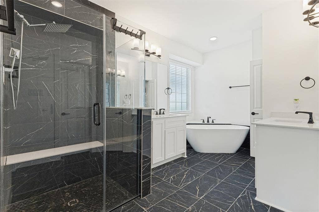 Walk-in shower featuring dark stone-finish tile, glass enclosure, and built-in bench, paired with a freestanding soaking tub, dual white vanity cabinets, and dark stone-finish floor tile