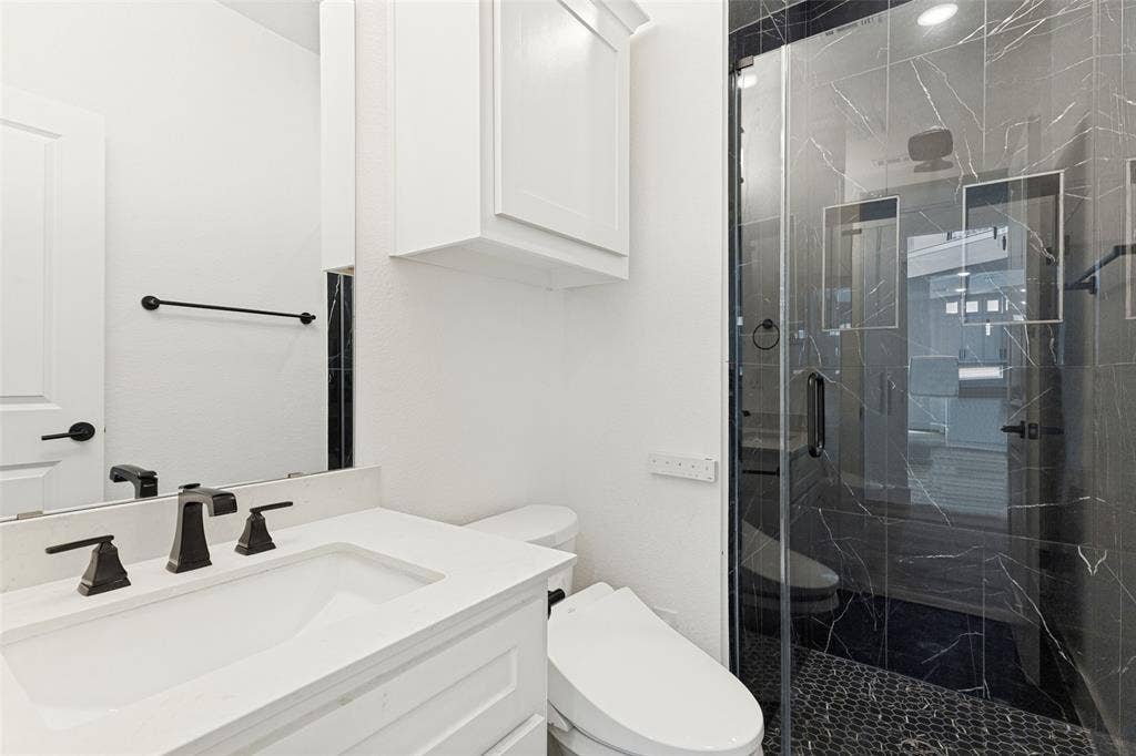 White vanity with a solid-surface countertop and integrated sink, matte black widespread faucet, walk-in shower with dark tile surround and pebble flooring, and built-in upper wall cabinetry