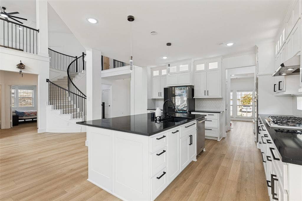 Kitchen featuring white cabinetry, a center island with a dark countertop and undermount sink, gas cooktop, and wood-finish flooring, complemented by a curved staircase with wrought iron railing