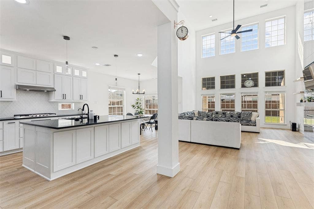 Open-concept kitchen and living area featuring high ceilings, white shaker-style cabinetry, dark stone countertops, wood-finish flooring, and multi-level windows