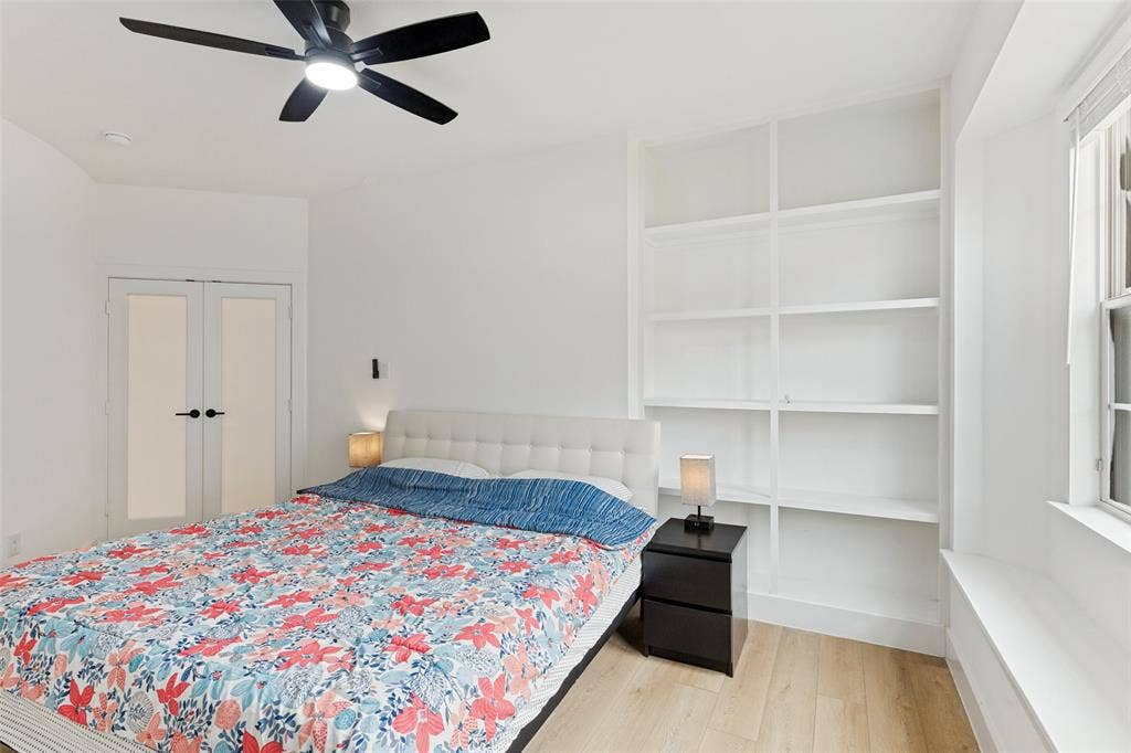 Room featuring light-toned wood-finish flooring, built-in white shelving, recessed window seat, matte black ceiling fan with integrated lighting, and double doors with frosted glass panes