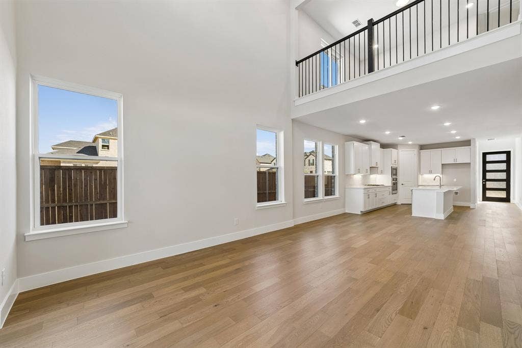 High-ceilinged living area featuring wood-finish flooring, an open-concept kitchen with white cabinetry and an island, and a second-level overlook with metal railings