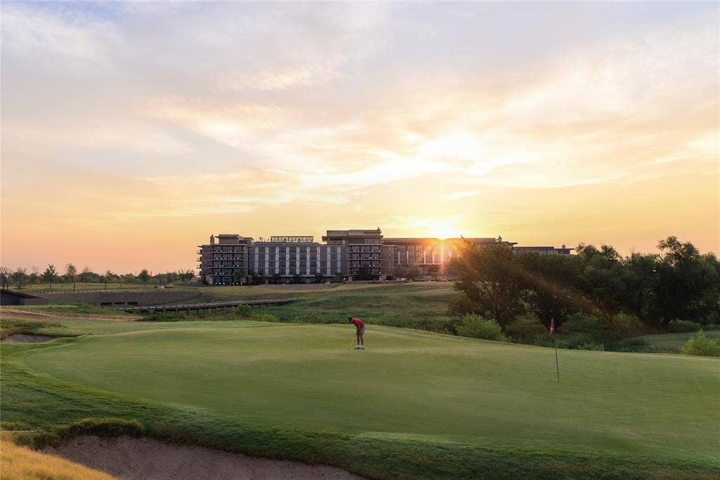 Golf course featuring a putting green, sand bunker, and flagstick, set against a backdrop of mature trees and a multi-story building