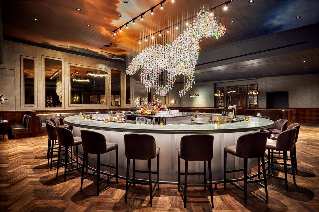 Circular bar featuring a translucent base, herringbone-patterned wood flooring, custom crystal light fixture, wall-mounted recessed mirrors with decorative trim, and an expansive cloud-patterned ceiling mural