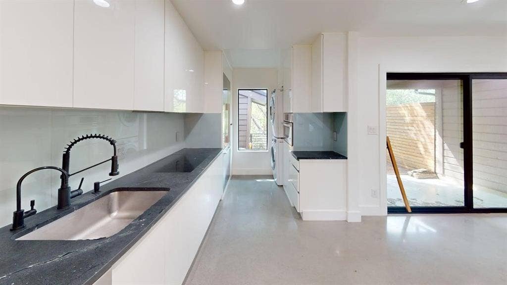 Modern kitchen featuring high-gloss white cabinetry, dark stone countertops, stainless steel undermount sink with a matte black arched faucet, and large-format sliding glass doors