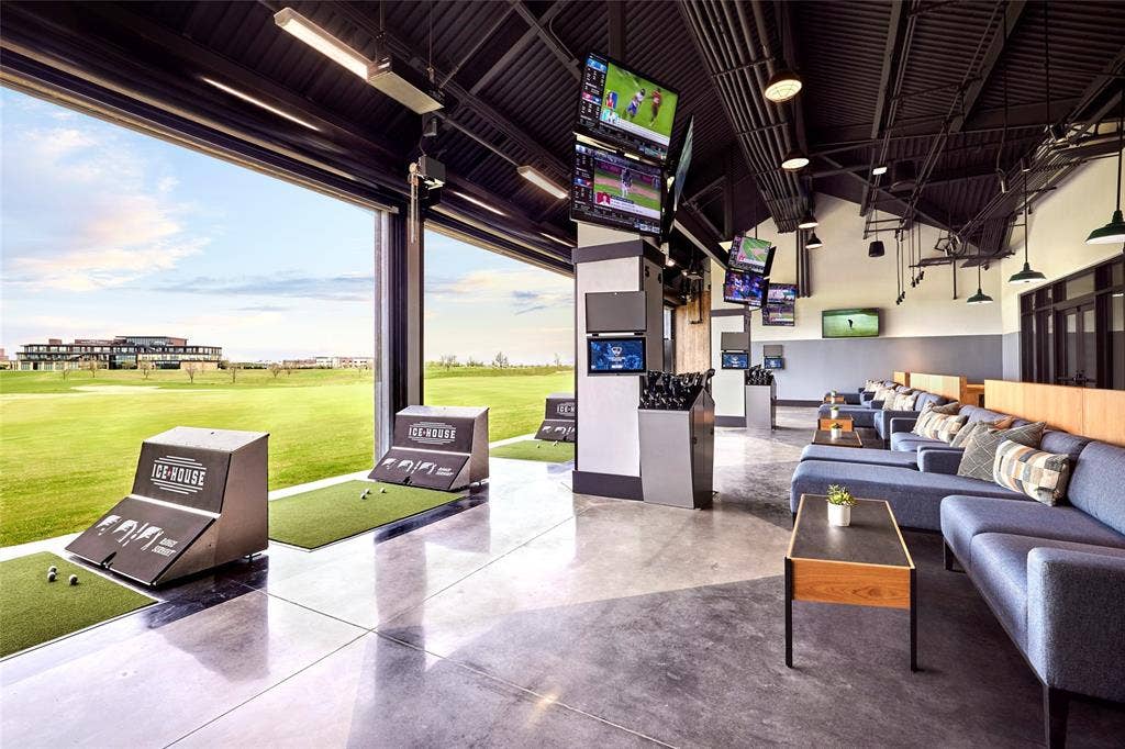 FRISCO PGA Expansive golf practice facility featuring high-ceiling structural beams, polished concrete flooring, integrated ceiling-mounted lighting, and an open-air wall design overlooking a grass course