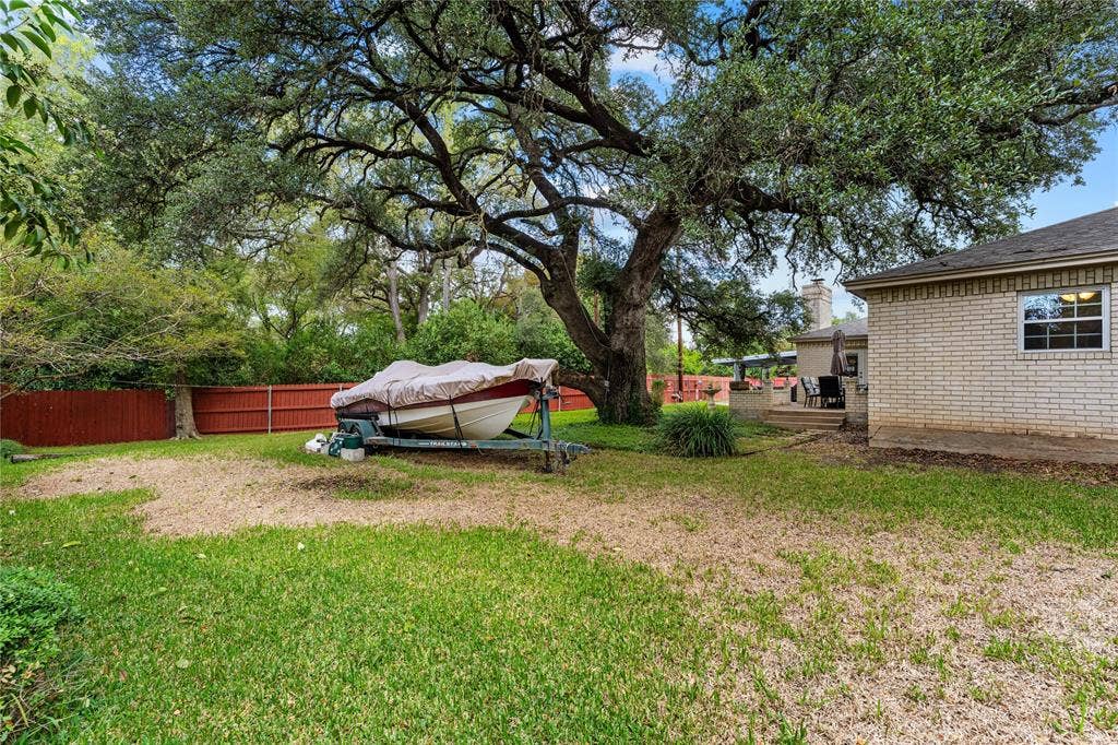 Backyard featuring a mature oak tree, light-colored brick exterior, red wood-panel privacy fencing, and a concrete patio