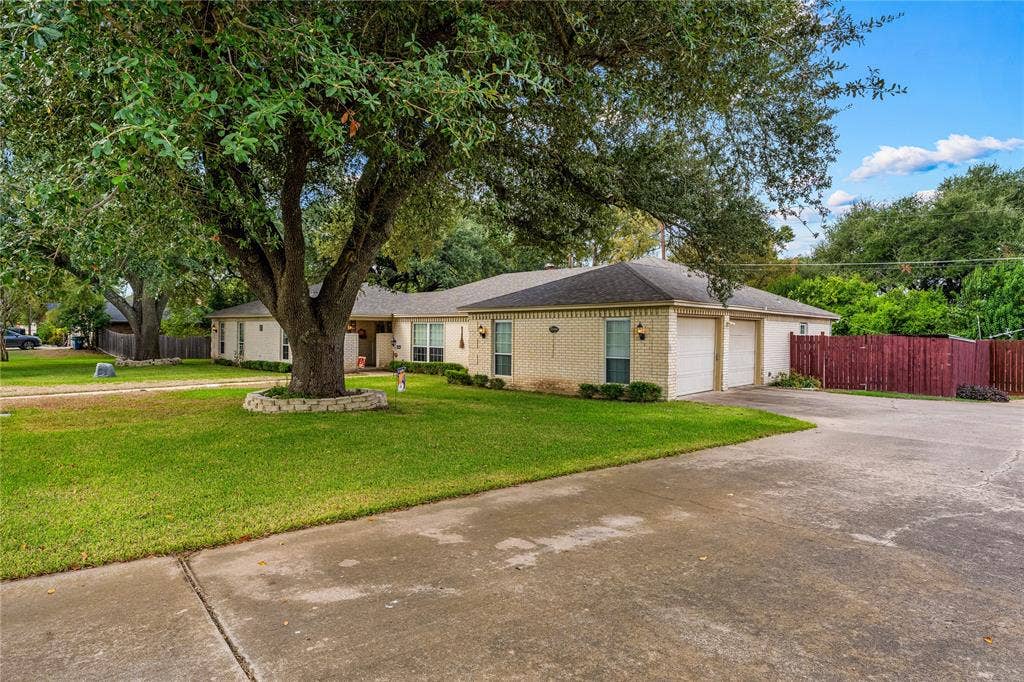 Single-story exterior featuring light-colored brick masonry, an attached two-car garage, concrete driveway, and mature landscaping with established trees