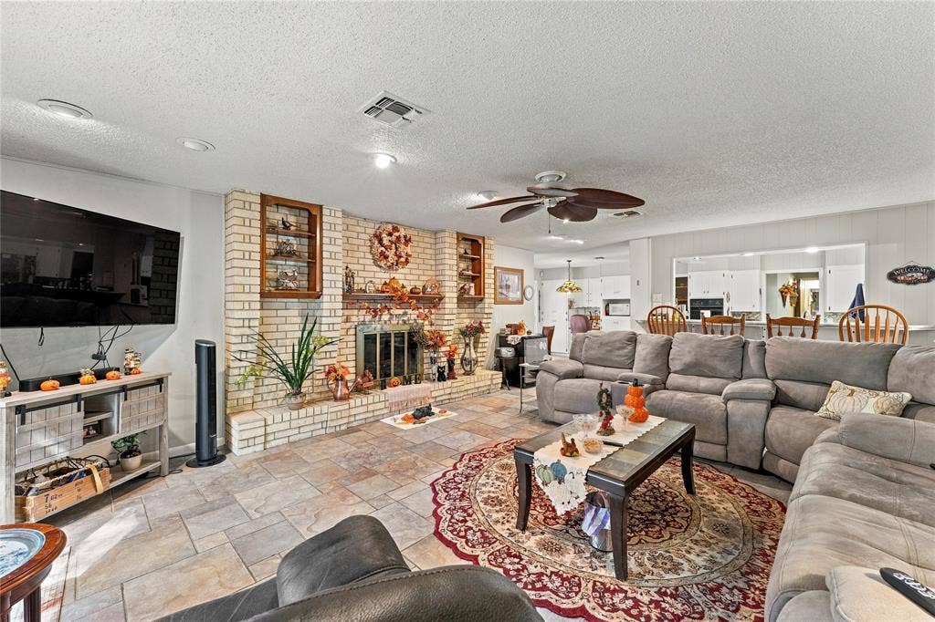 Floor-to-ceiling light-colored brick fireplace with built-in display shelving, stone-finish tile flooring, ceiling fan, and open-concept pass-through window
