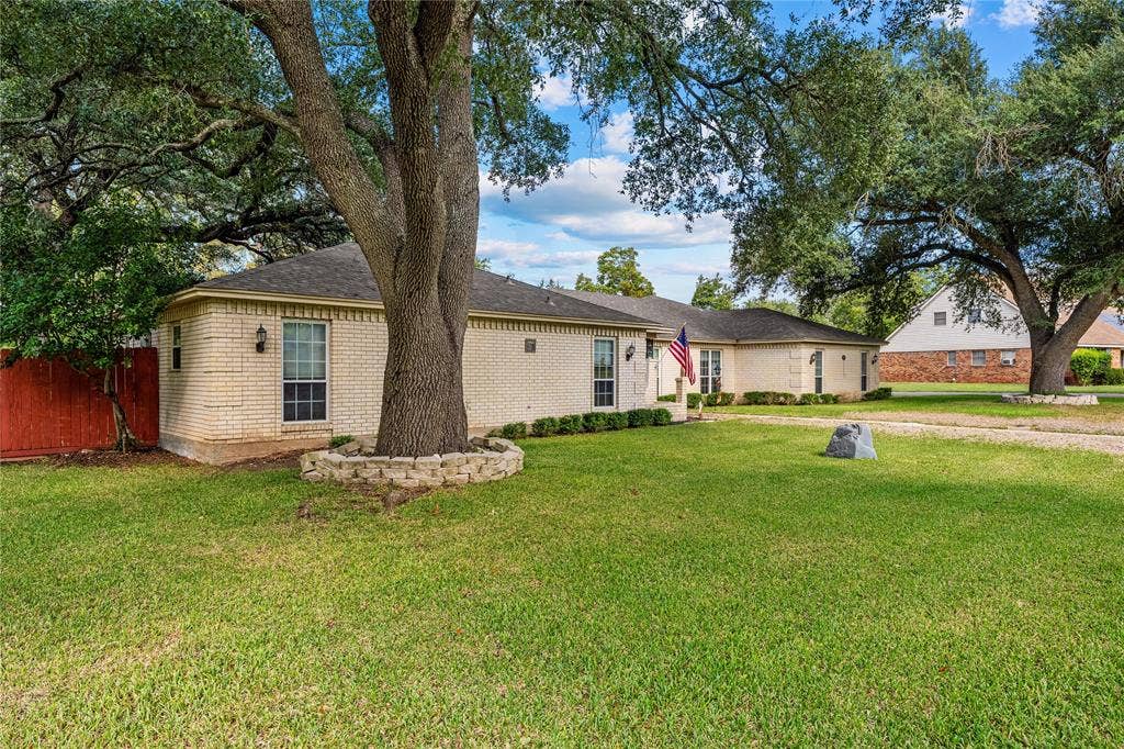 Light-colored brick exterior featuring a stone-lined tree planter, mature canopy trees, a sprawling lawn, and an attached red privacy fence