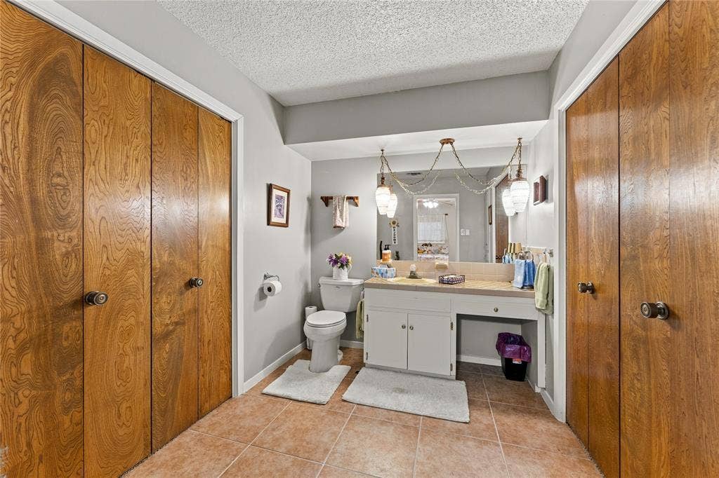 Bathroom featuring ceramic tile flooring, dual floor-to-ceiling wood-finish closet doors, a white vanity with a neutral-tone countertop, and a full-width mirror with decorative pendant lighting
