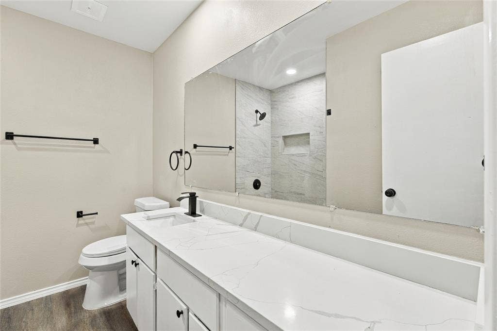 Bathroom featuring a dual-sink vanity with a solid-surface countertop, full-width wall mirror, wood-finish flooring, and a walk-in shower with stone-style wall tile and an integrated recessed niche