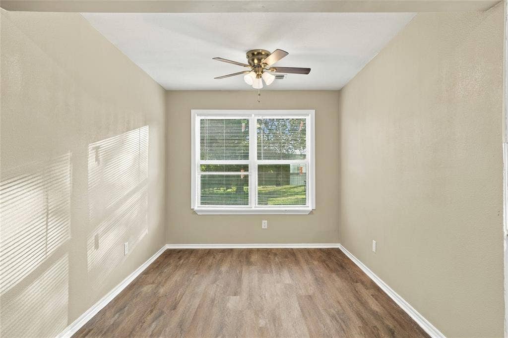 Neutral-toned interior space featuring wood-finish flooring, a multi-light ceiling fan, and a large window with white trim