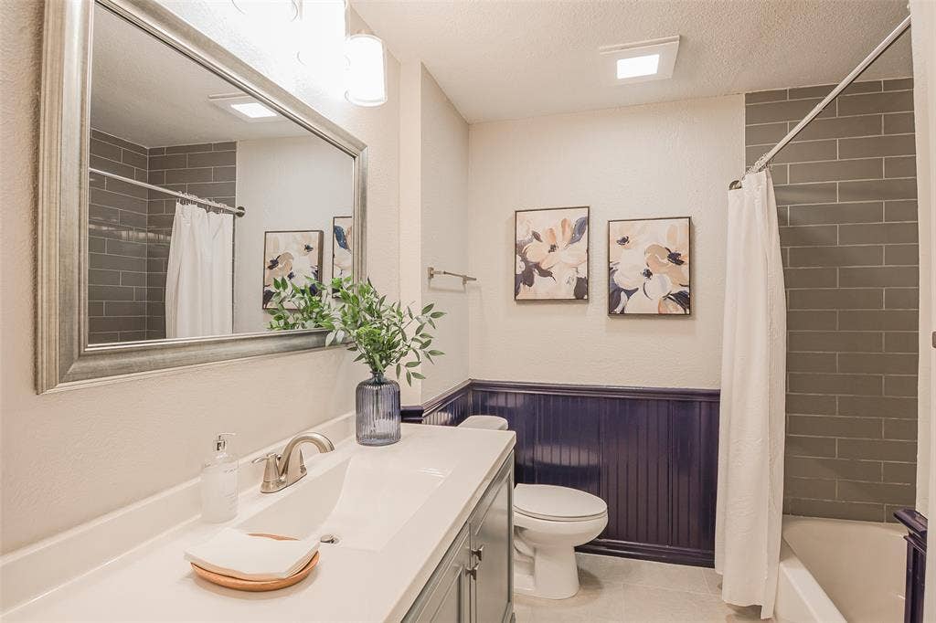 8050 Fall River Drive, Dallas, TX 75228 Bathroom featuring a vanity with an integrated basin, a large framed mirror, dark wood-tone wainscoting, and a tiled shower surround