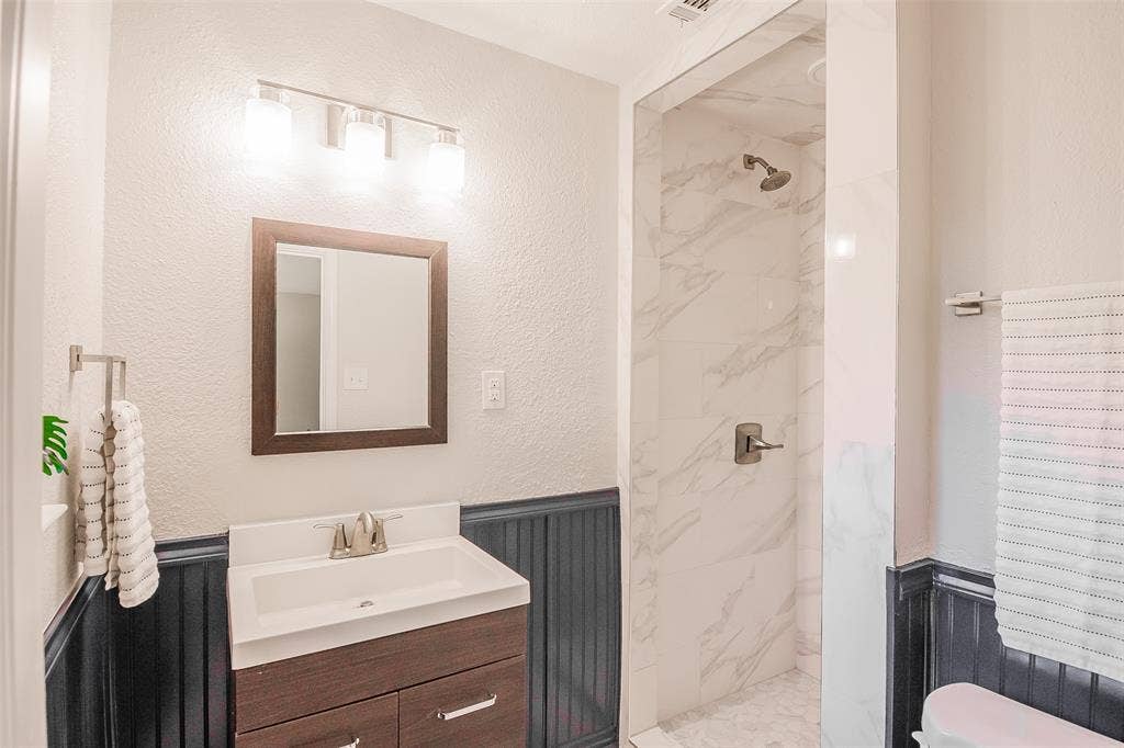 8050 Fall River Drive, Dallas, TX 75228 Bathroom featuring a vanity with a white rectangular sink, dark wood-finish cabinetry, wainscot paneling, a shower enclosure with stone-patterned tile, and a three-light wall-mounted fixture
