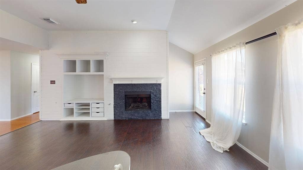 Living area featuring wood-finish flooring, a vaulted ceiling, a floor-to-ceiling painted brick fireplace with a textured stone surround, and built-in shelving with integrated storage drawers