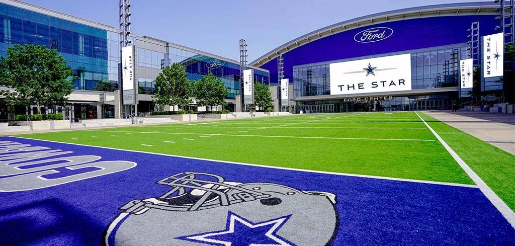 The Star District Synthetic turf athletic field featuring professional markings, paved pedestrian walkways, expansive glass-facade exterior structures, and integrated stadium lighting