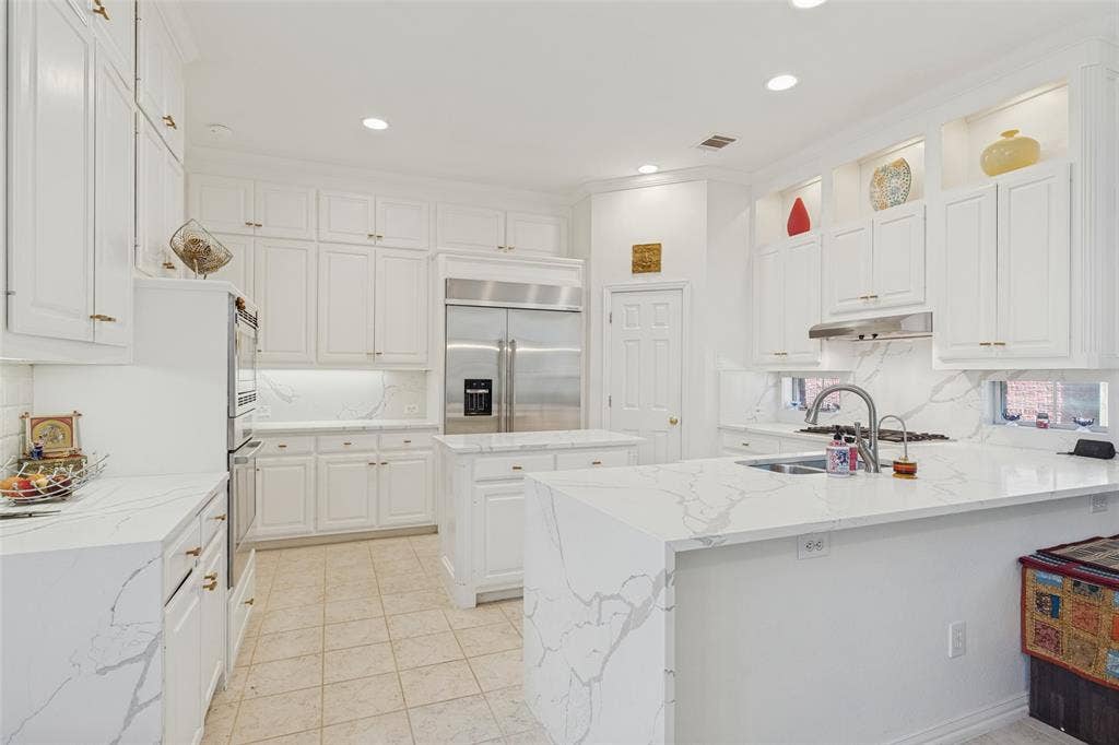 Kitchen featuring custom white cabinetry, stone-surface countertops with waterfall edge, built-in stainless steel refrigerator, recessed lighting, and integrated glass-front display shelving