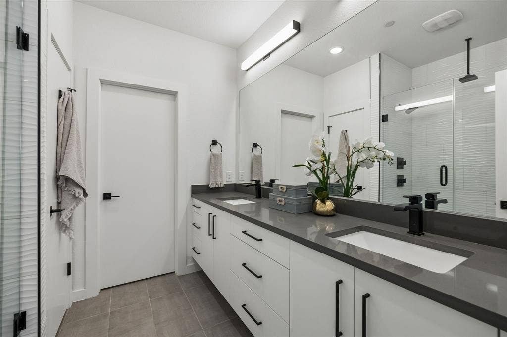 1901 N Carroll Avenue, #104, Dallas, TX 75204 Dual-sink vanity with matte dark hardware, charcoal-toned solid surface countertop, expansive wall-to-wall mirror, tiled flooring, and walk-in shower with glass enclosure