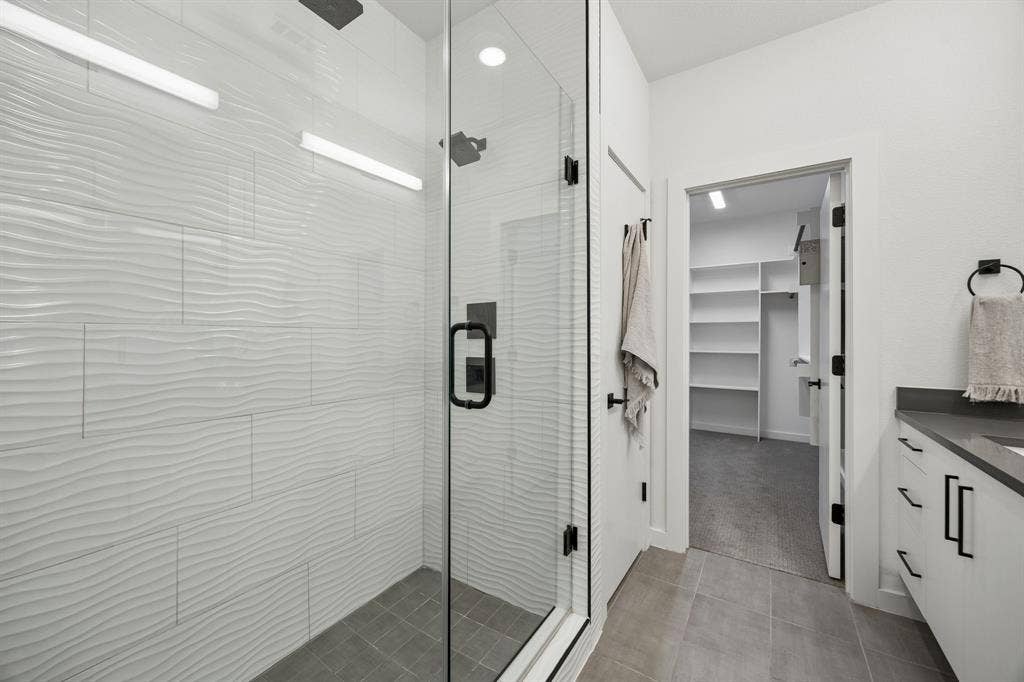 1901 N Carroll Avenue, #104, Dallas, TX 75204 Walk-in shower featuring textured white tile surround, glass enclosure, matte black fixtures, and grey floor tiling, complemented by a white vanity with dark countertop and built-in shelving