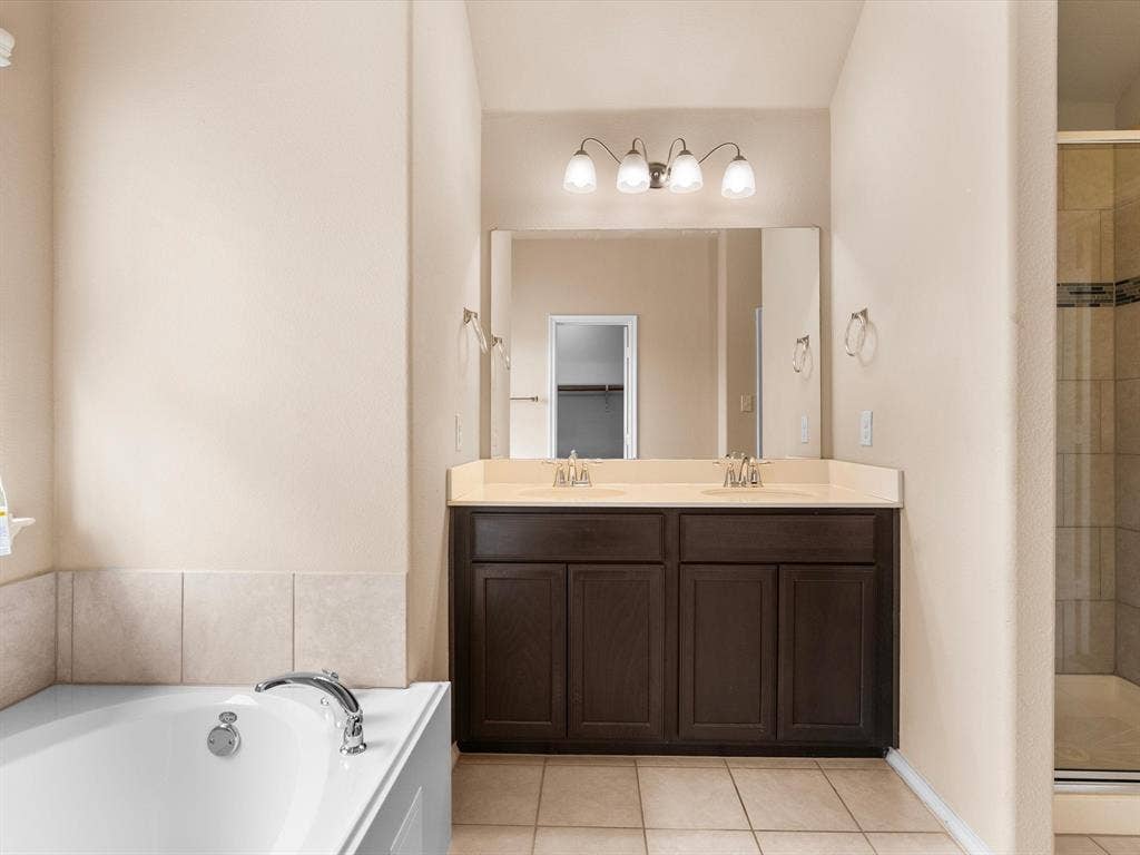 Dark wood dual-sink vanity with solid-surface countertop, large wall-mounted mirror, soaking tub, glass-enclosed shower, and tile flooring