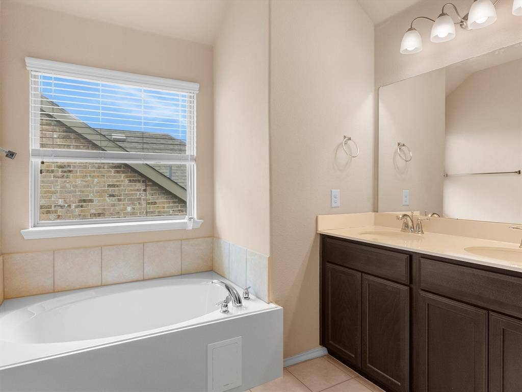 Built-in soaking tub with a tiled surround, dual-sink vanity featuring dark-toned cabinetry, solid surface countertop, wall-to-wall mirror, and three-light vanity fixture