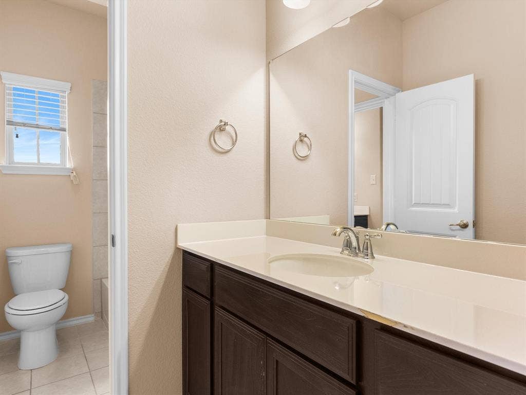 Bathroom featuring dark wood-finish cabinetry, solid-surface countertop with an integrated sink, full-width wall mirror, and neutral tile flooring