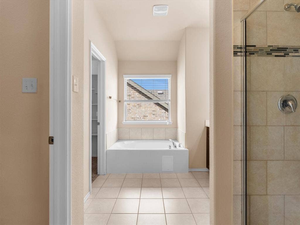 Soaking tub with tiled surround, walk-in glass-enclosed shower featuring decorative tile inlay, and neutral floor tile