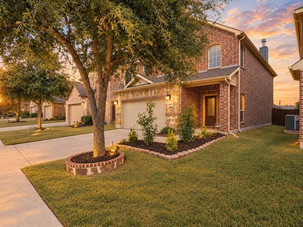 Two-story brick exterior featuring stone masonry accents, an arched window, a concrete driveway, and landscaped garden beds with decorative stone edging