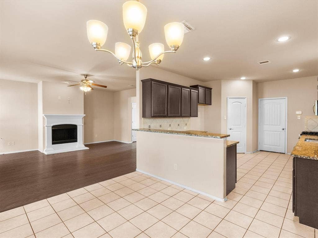 Kitchen featuring dark cabinetry, stone-finish countertops, tile flooring, and a multi-arm chandelier, alongside a living area with a fireplace and wood-finish flooring