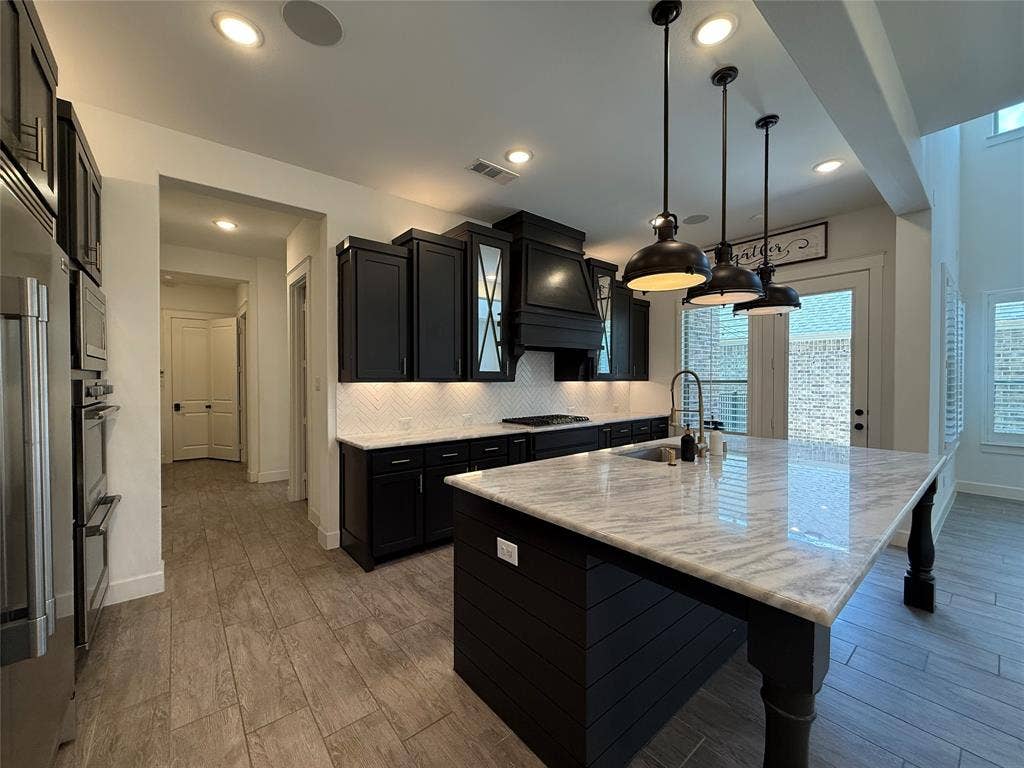 12905 Yale Court, Frisco, TX 75035 Kitchen featuring dark cabinetry, expansive stone-topped island with seating, herringbone tile backsplash, stainless steel built-in double ovens, and suspended industrial-style pendant lighting