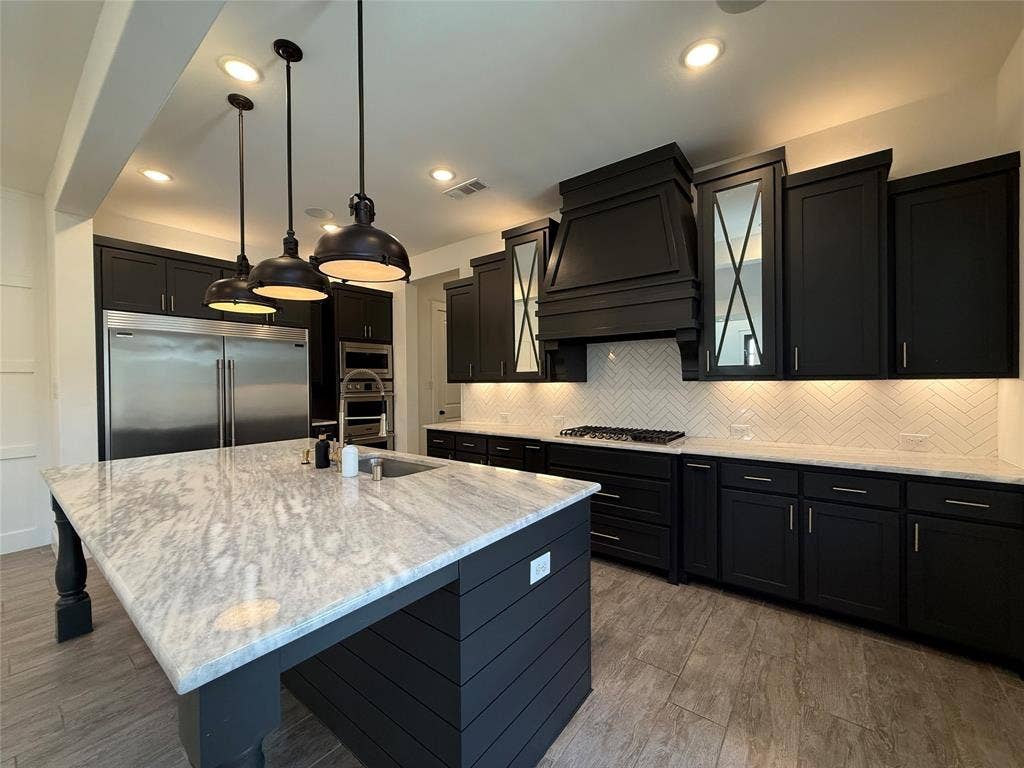 12905 Yale Court, Frisco, TX 75035 Kitchen featuring dark cabinetry, expansive stone-topped island with integrated seating, stainless steel appliances, herringbone tile backsplash, and oversized metal pendant lighting