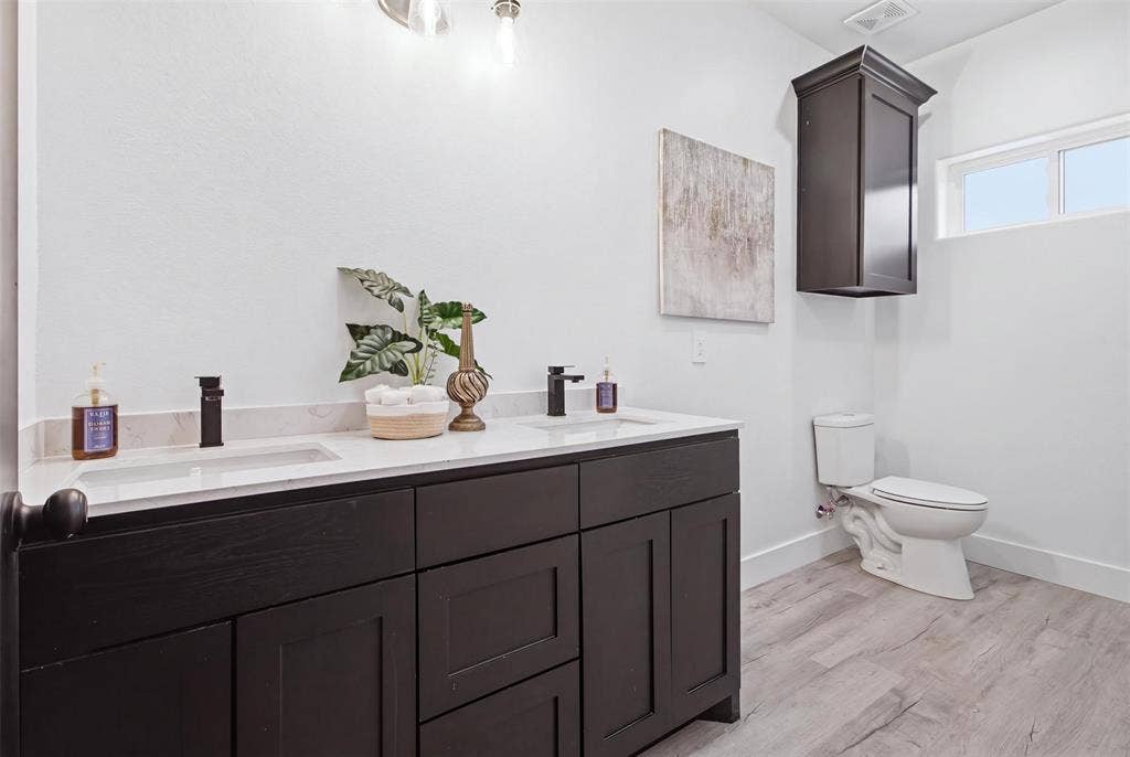 Double vanity featuring dark-toned cabinetry, light-colored stone countertops with integrated rectangular sinks, matte black faucets, and wood-finish flooring, complemented by an overhead storage cabinet