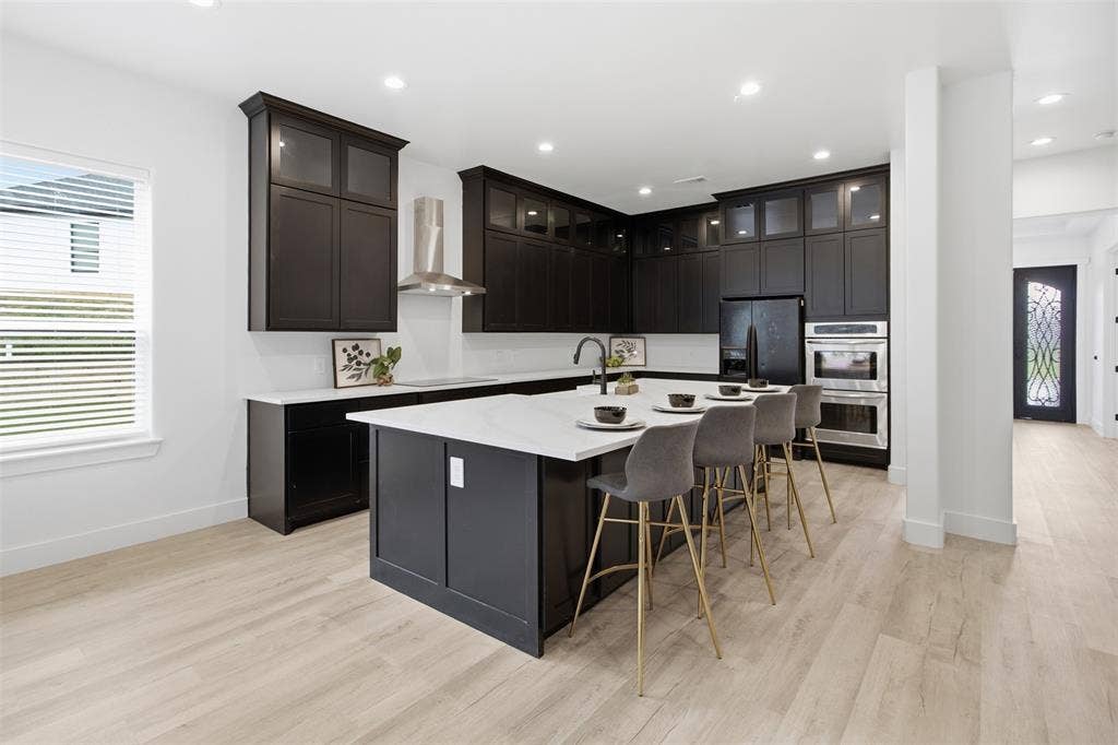 Kitchen featuring dark cabinetry with glass-fronted upper units, a large central island with a stone-surface countertop, integrated stainless steel double wall ovens, a stainless steel range hood, and light-toned wood-finish flooring