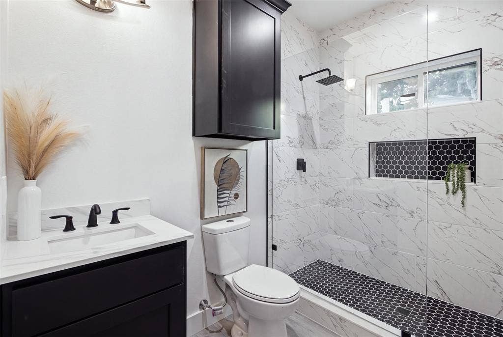 Bathroom featuring a dark wood-finish vanity with a stone countertop, wall-mounted matte black cabinetry, and a walk-in shower with floor-to-ceiling veined tile, contrasting hexagonal tile flooring, and matte black fixtures