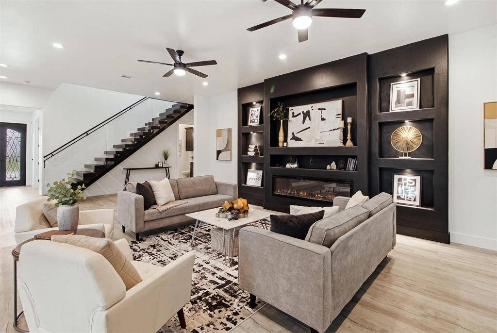 Floor-to-ceiling black feature wall with integrated shelving and fireplace, wood-finish flooring, staircase with glass railing, and ceiling fans