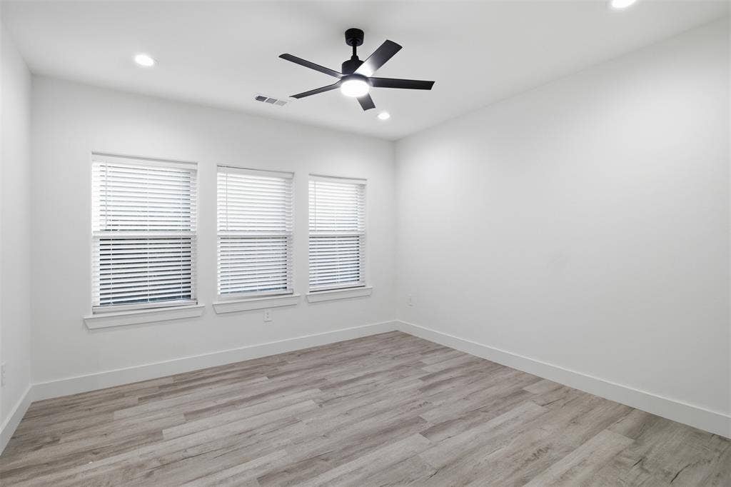Neutral-toned room featuring wood-finish flooring, three windows with horizontal blinds, and a ceiling-mounted light fixture with a fan