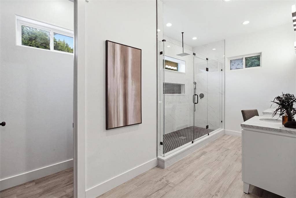 Walk-in shower with a glass enclosure, rainfall showerhead, and mosaic tile flooring, complemented by a vanity with a stone-finish countertop and wood-finish flooring