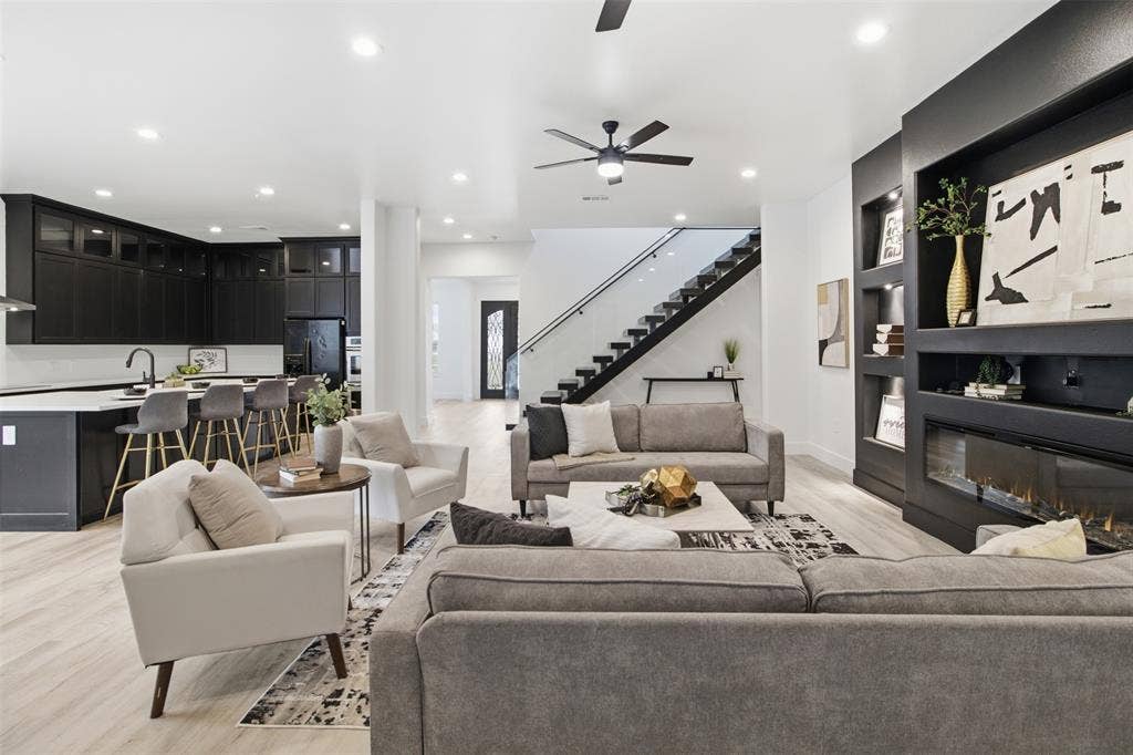 Open-concept living space featuring wood-finish flooring, a kitchen with dark cabinetry and a central island, a built-in wall unit with an integrated linear fireplace, and a staircase with a glass-panel railing