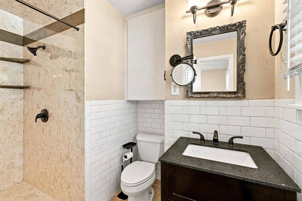 Bathroom featuring a shower with stone-finish tile surround and built-in shelving, dark vanity with a rectangular basin, white subway tile wainscoting, and wall-mounted lighting fixtures