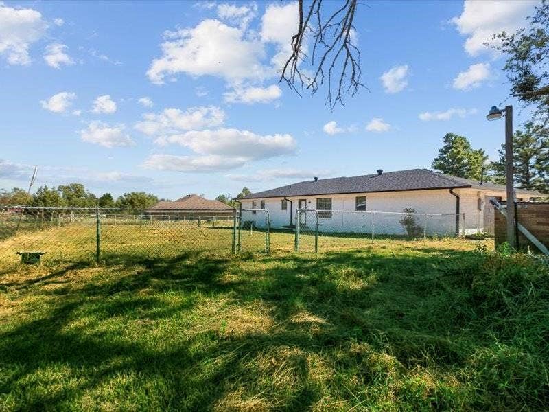 17014 Woodcrest Circle, Lindale, TX 75771 Exterior rear elevation featuring a light-colored masonry facade, a dark-toned shingled roof, a chain-link fenced perimeter, and an open grass lot