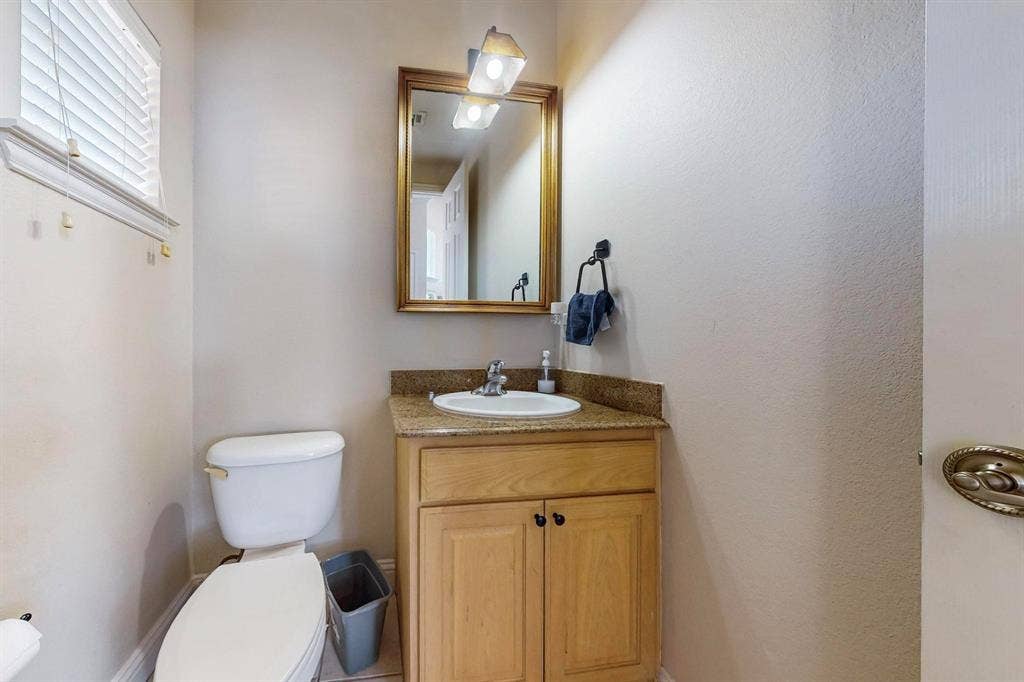2606 Rue De Ville, Irving, TX 75038 Vanity featuring light wood-finish cabinetry, a stone-composite countertop with an integrated drop-in sink, a wall-mounted light fixture, and a framed mirror