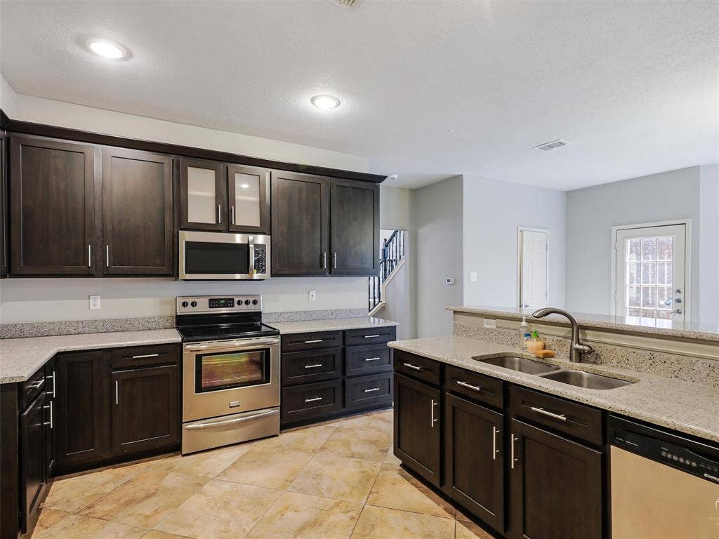 Kitchen featuring dark wood cabinetry, speckled stone countertops, stainless steel appliances, a dual-basin undermount sink with a gooseneck faucet, and tiled flooring