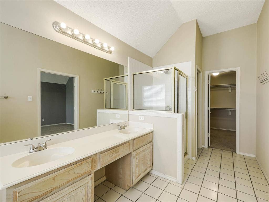 Dual-sink vanity with light wood-finish cabinetry, full-width wall mirror, tile flooring, glass-enclosed shower, and built-in closet shelving