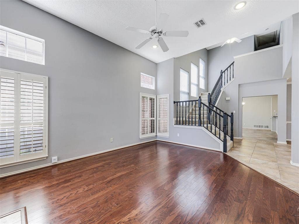 High-ceilinged living area featuring wood-finish flooring, plantation shutters, a multi-blade ceiling fan, and a staircase with decorative wrought iron railing