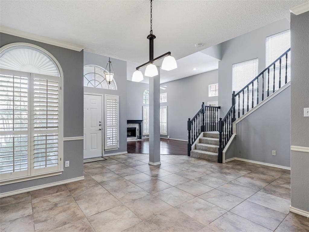 High-ceiling interior featuring tile flooring, arched windows with plantation shutters, a multi-light chandelier, and a staircase with a dark-finished handrail and balusters