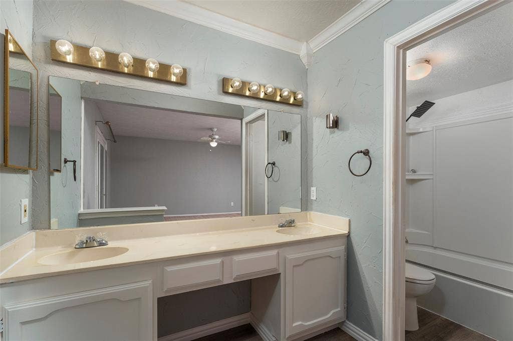 826 Camino Court, Grand Prairie, TX 75052 Dual-sink vanity with a countertop, expansive wall-to-wall mirror, double multi-bulb light fixtures, crown molding, and a tub-shower combination unit