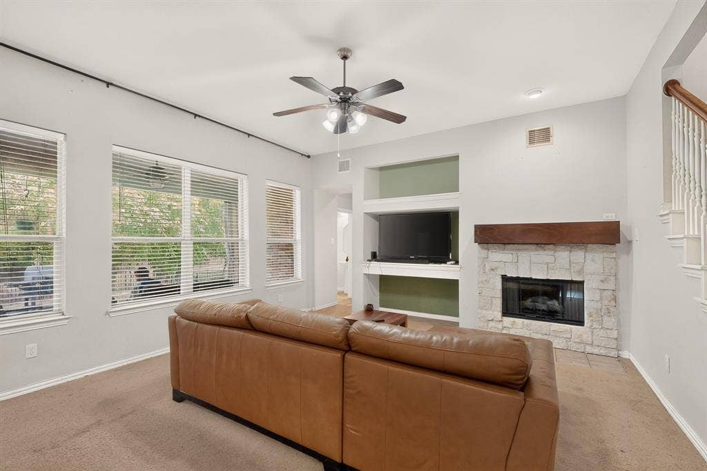 Living area featuring a floor-to-ceiling stone fireplace with a wood mantel, built-in shelving, multiple windows with horizontal blinds, and a ceiling fan