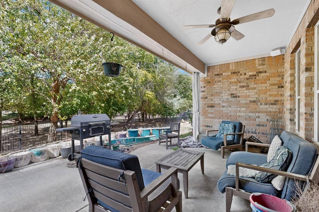 Covered concrete patio featuring a brick accent wall, ceiling fan with integrated lighting, and a metal perimeter fence